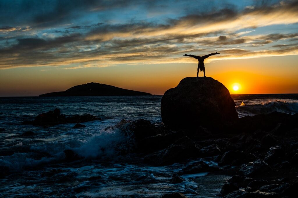 Vicki auf einem Halbrunden Felsen im Handstand stehend. Hinten Meer und ein Sonnenuntergang mit Wolken